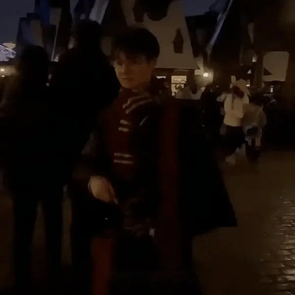 A person standing outdoors at night wearing a dark outfit with a red and black striped scarf and a dark coat draped over one shoulder. The background is a dimly lit public walkway with blurred figures and buildings.